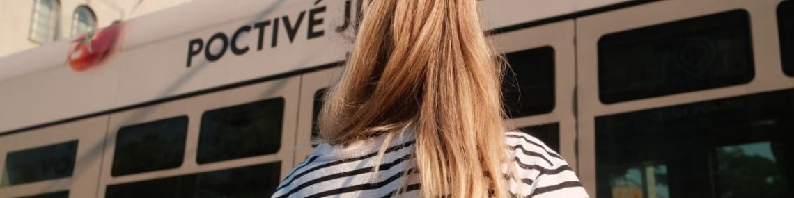 A woman with blonde hair in a striped shirt stands near a city tram on a sunny day.